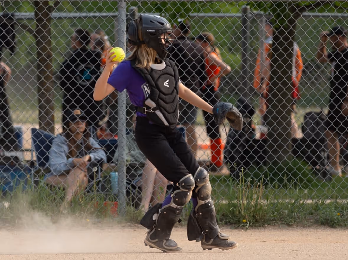 Young softball catcher in black protective gear and purple uniform preparing to throw a yellow softball on a dusty field.