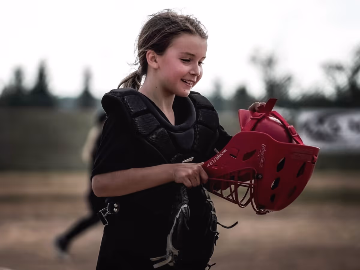 Young girl wearing black catcher's gear holding a red catcher's helmet and smiling on a baseball field.