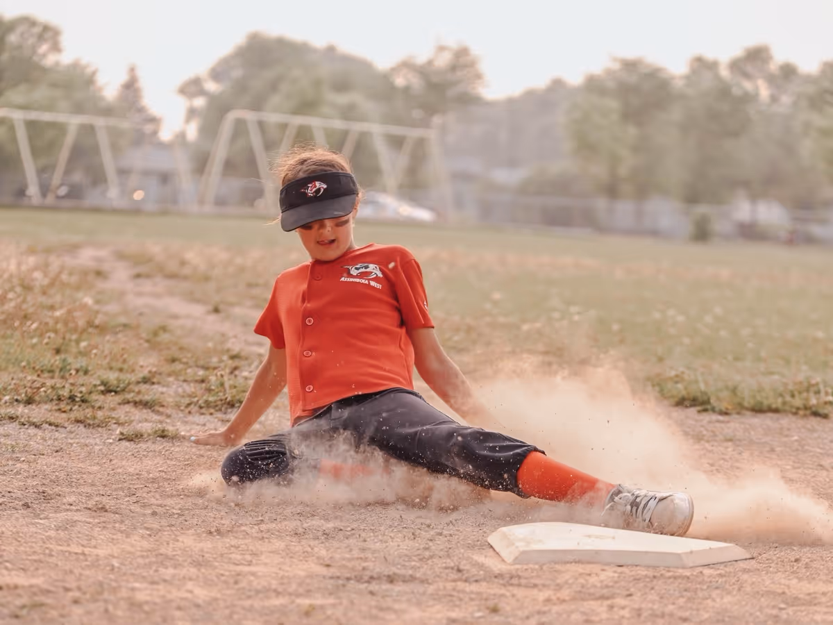 Youth baseball player in red jersey and black pants sliding into a base with dust flying around.