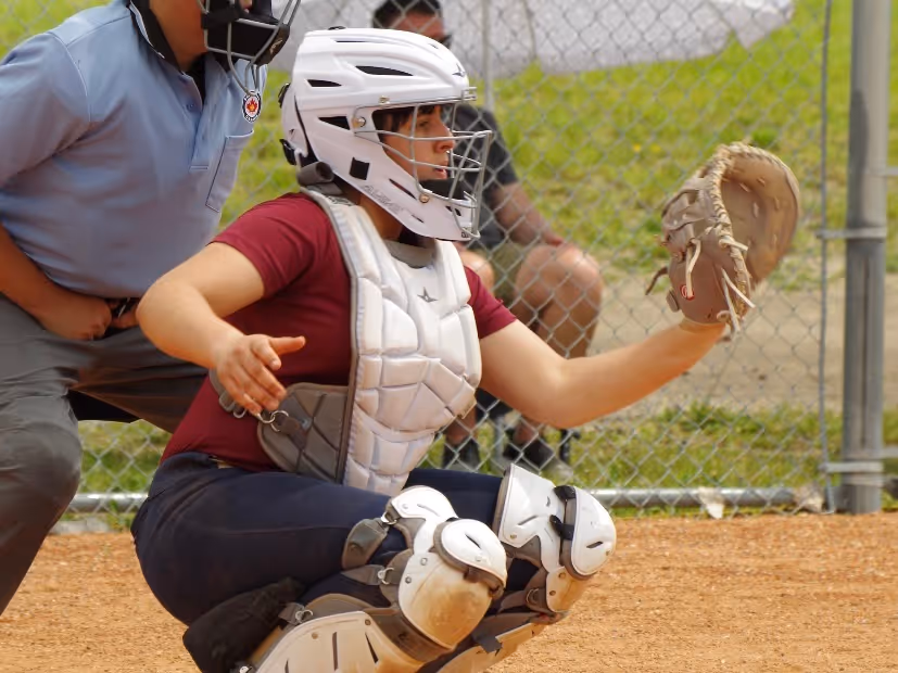 Female baseball catcher in white helmet and chest protector, crouching with glove extended ready to catch a ball.
