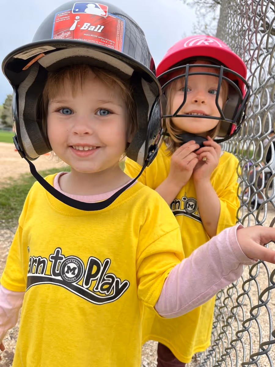 Two young children wearing yellow Learn to Play shirts and baseball helmets standing by a chain-link fence at a baseball field.