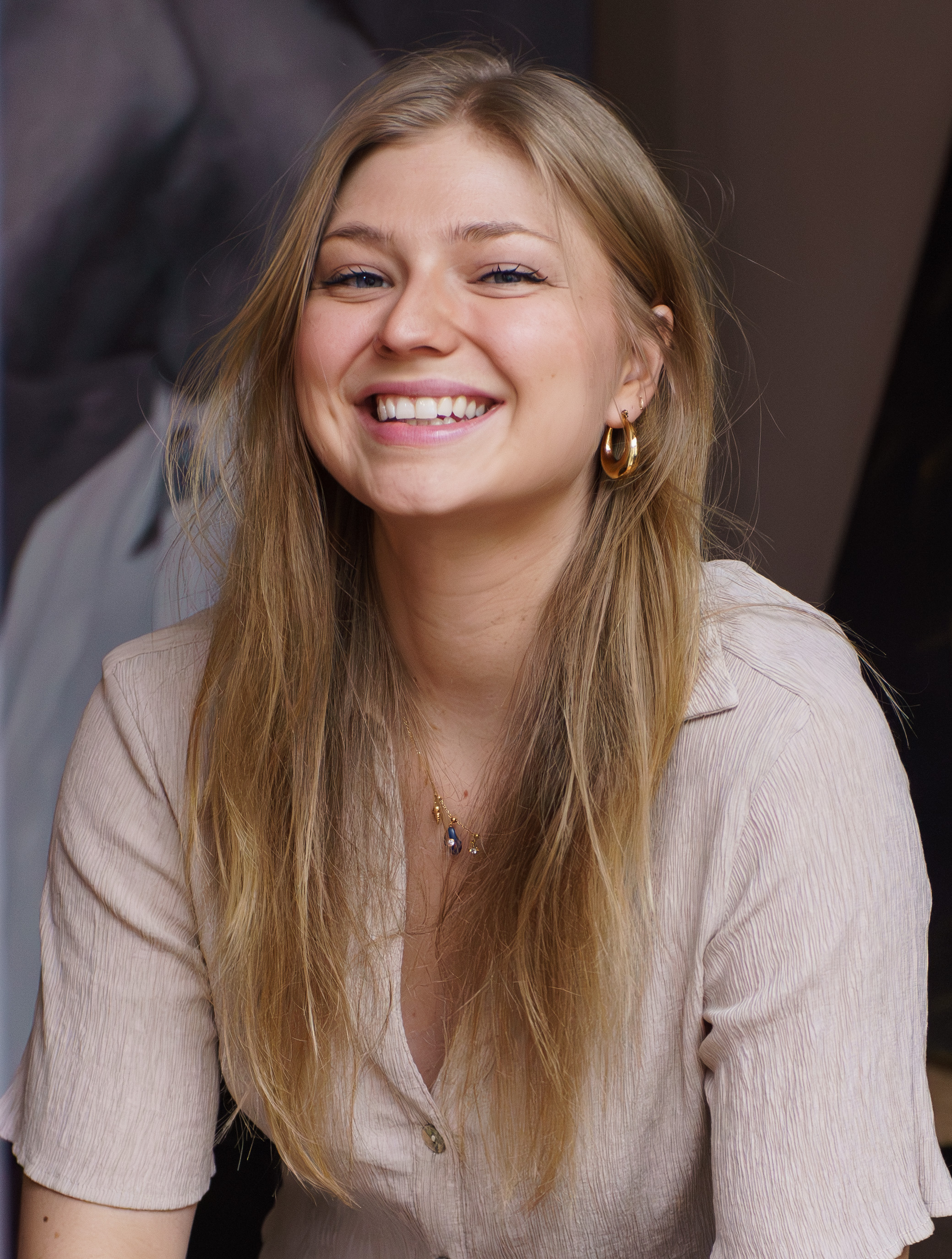 Smiling young woman with long blonde hair wearing gold hoop earrings and a light shirt.