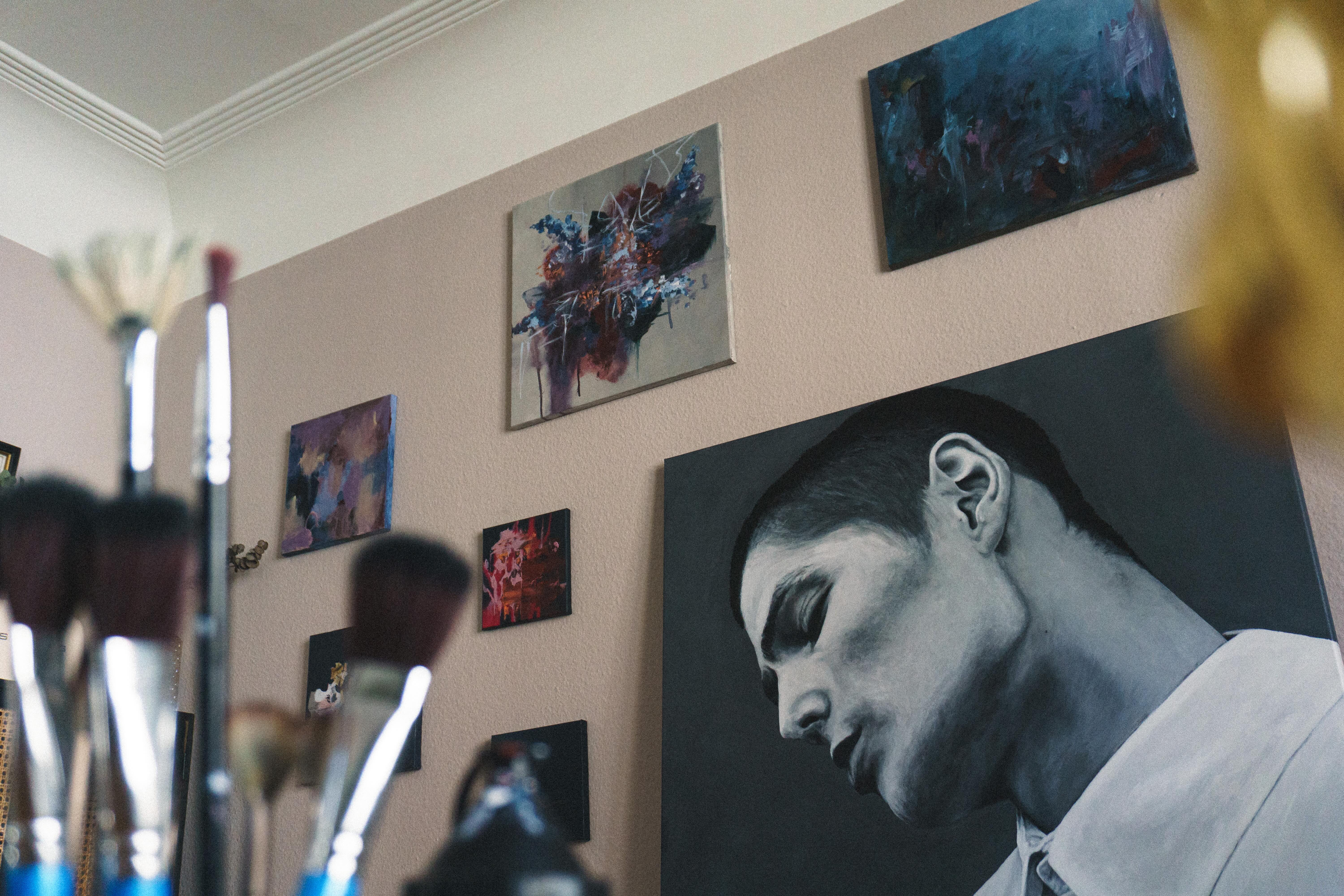 Abstract paintings and a monochrome portrait of a man with closed eyes on a beige wall.