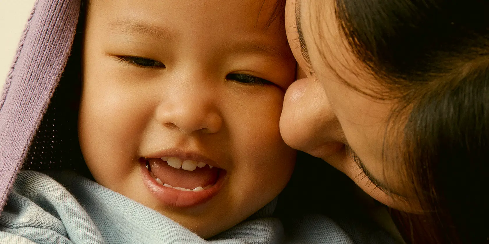 Close-up of a woman gently kissing a smiling young child wrapped in a purple blanket.