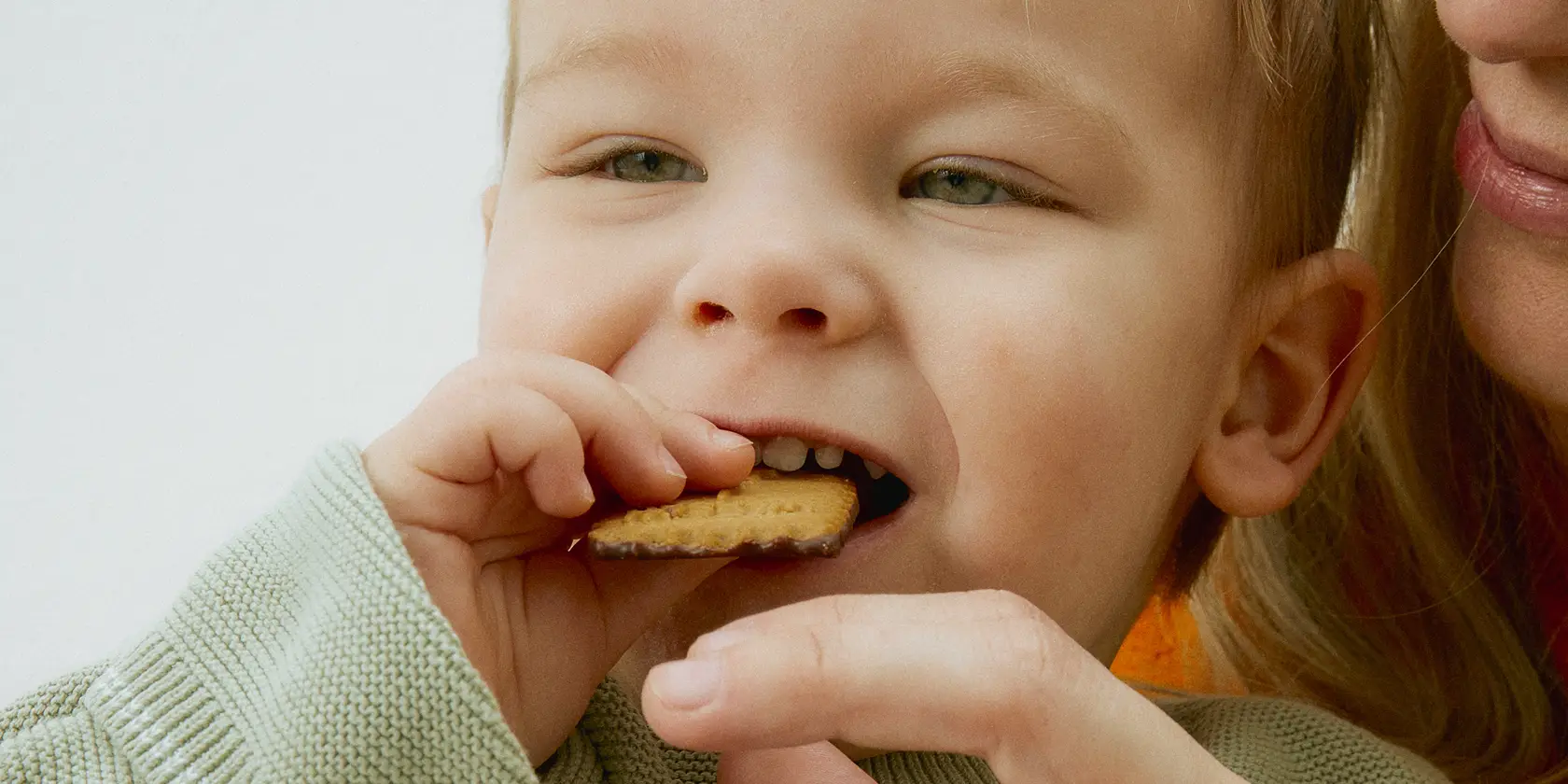 Close-up of a young child eating a biscuit with an adult gently supporting their hand.