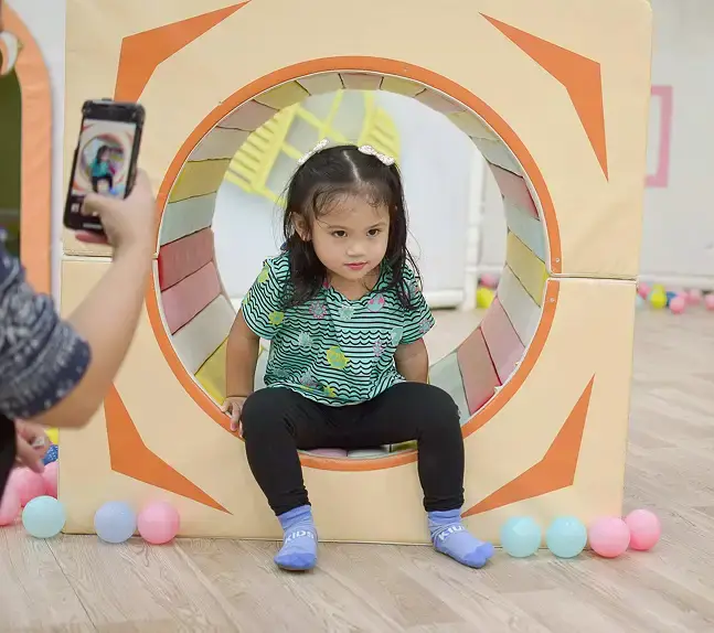Young girl crawling through a soft play tunnel while being photographed on a smartphone.