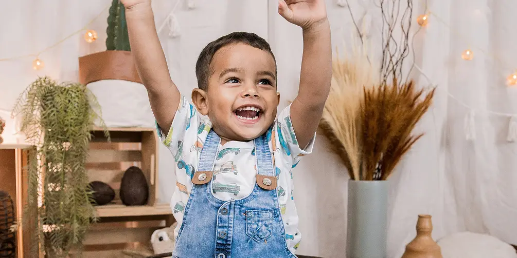 Smiling young child wearing blue denim overalls and a printed shirt, raising arms joyfully indoors with plants in the background.