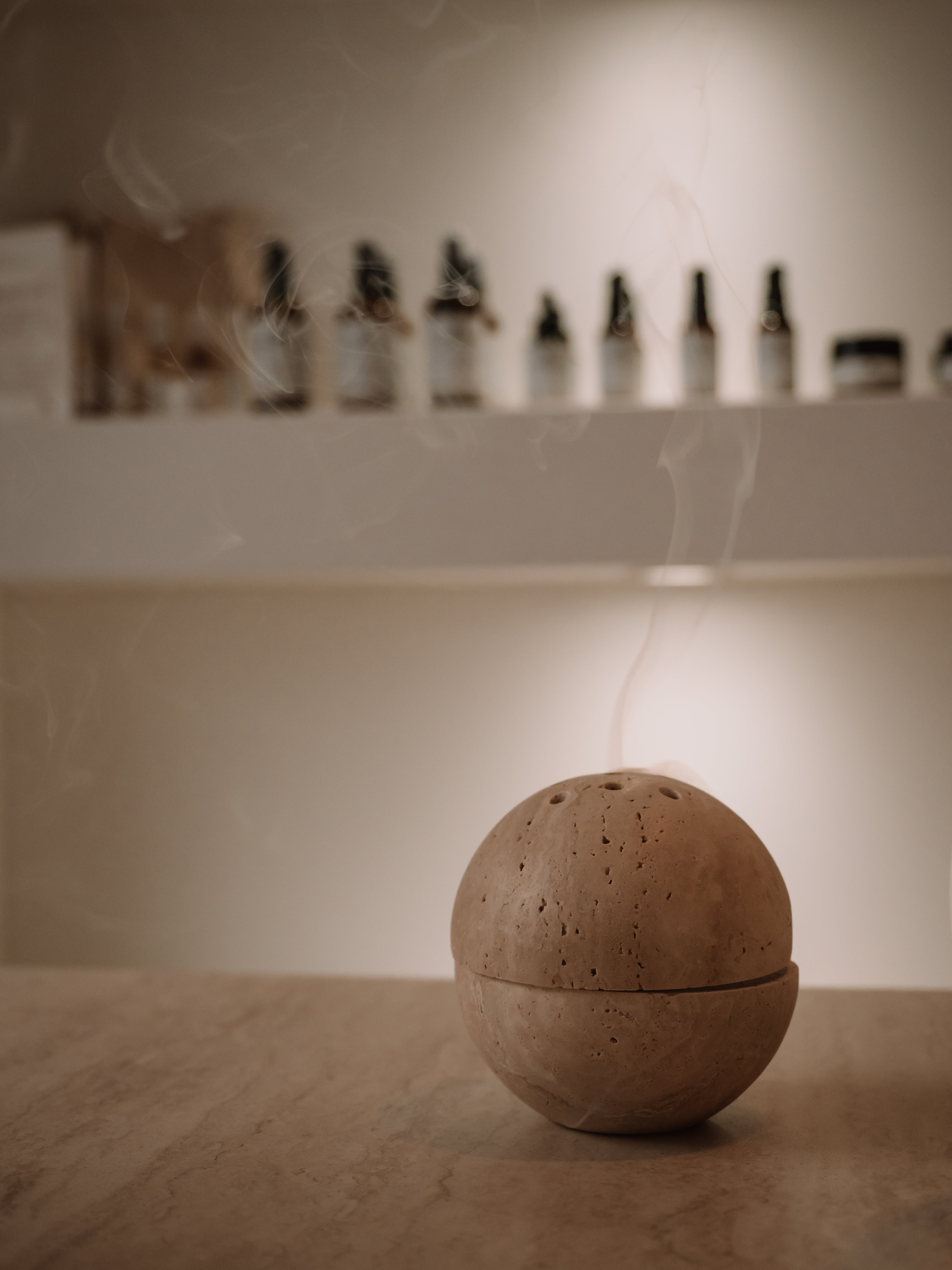 Round stone incense burner emitting smoke on a marble surface with blurred essential oil bottles in the background.