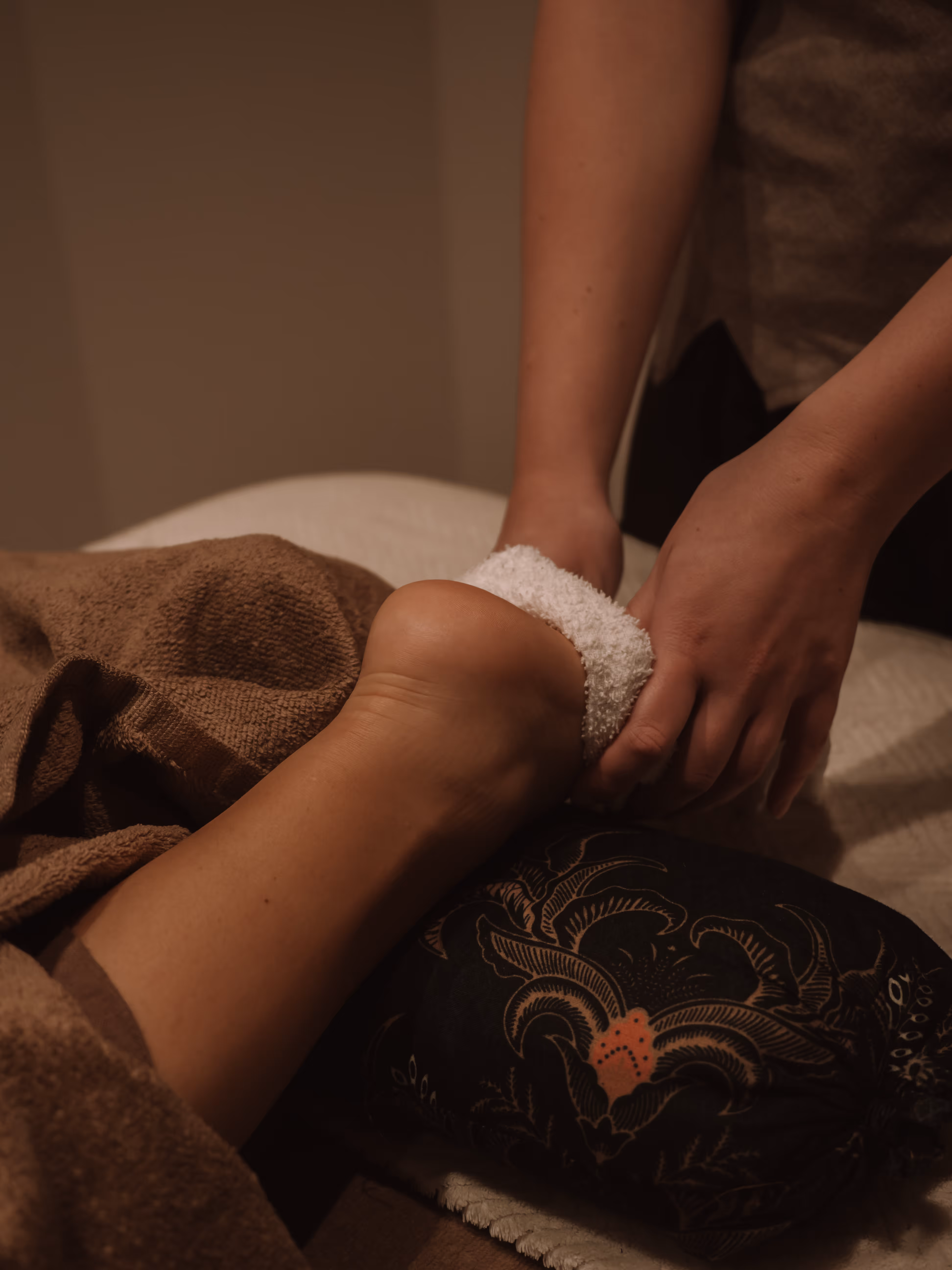 Person receiving a foot massage with a white towel wrapped around the heel and a patterned cushion under the ankle.