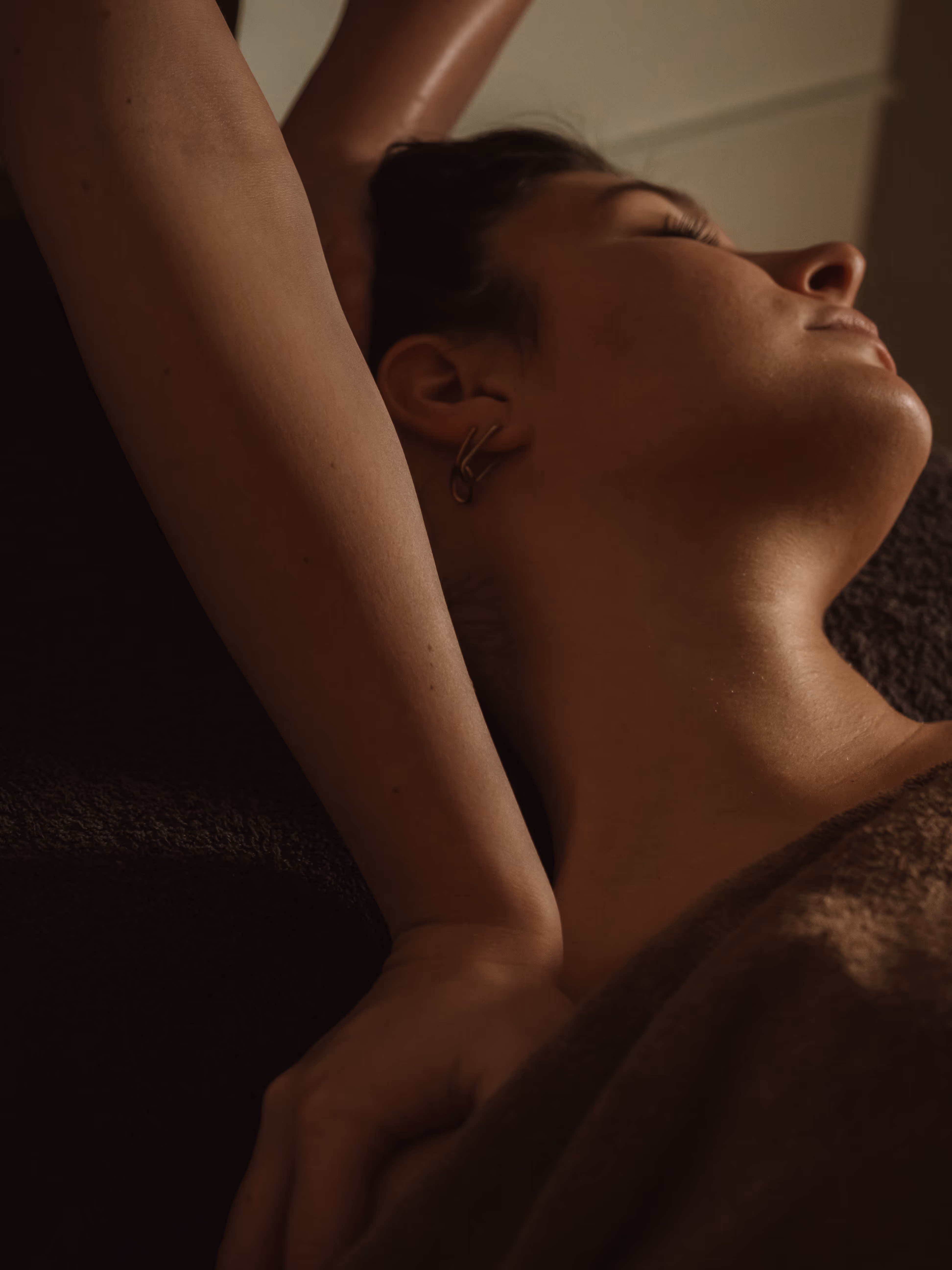 Close-up of a relaxed woman receiving a shoulder massage with her eyes closed and a brown blanket covering her.
