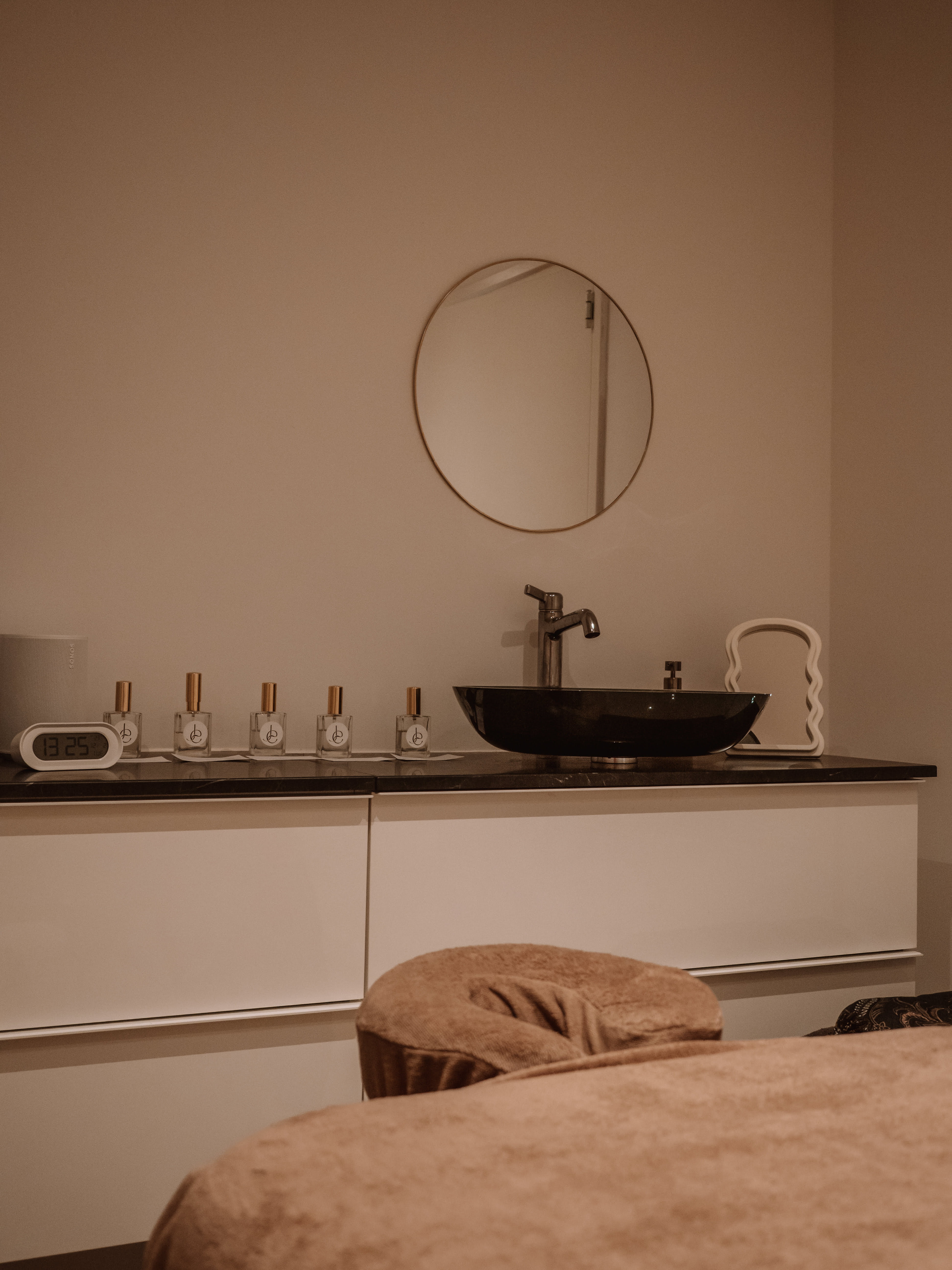 Minimalist treatment room with a brown massage table, round mirror, black sink, and five small bottles lined up on a countertop.