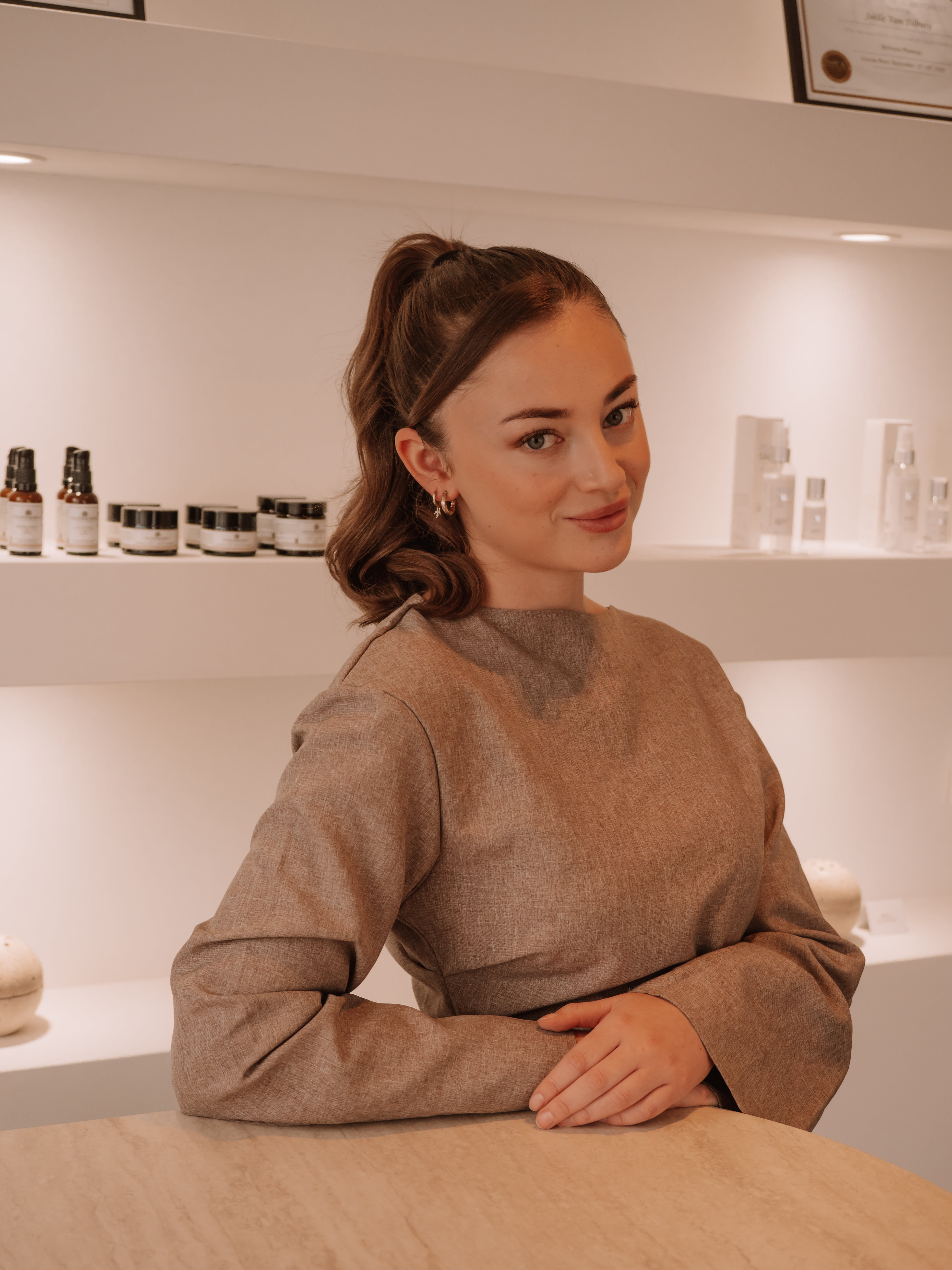 Young woman with brown hair tied in a ponytail, wearing a beige long-sleeve top, sitting at a table with skincare products displayed on shelves behind her.