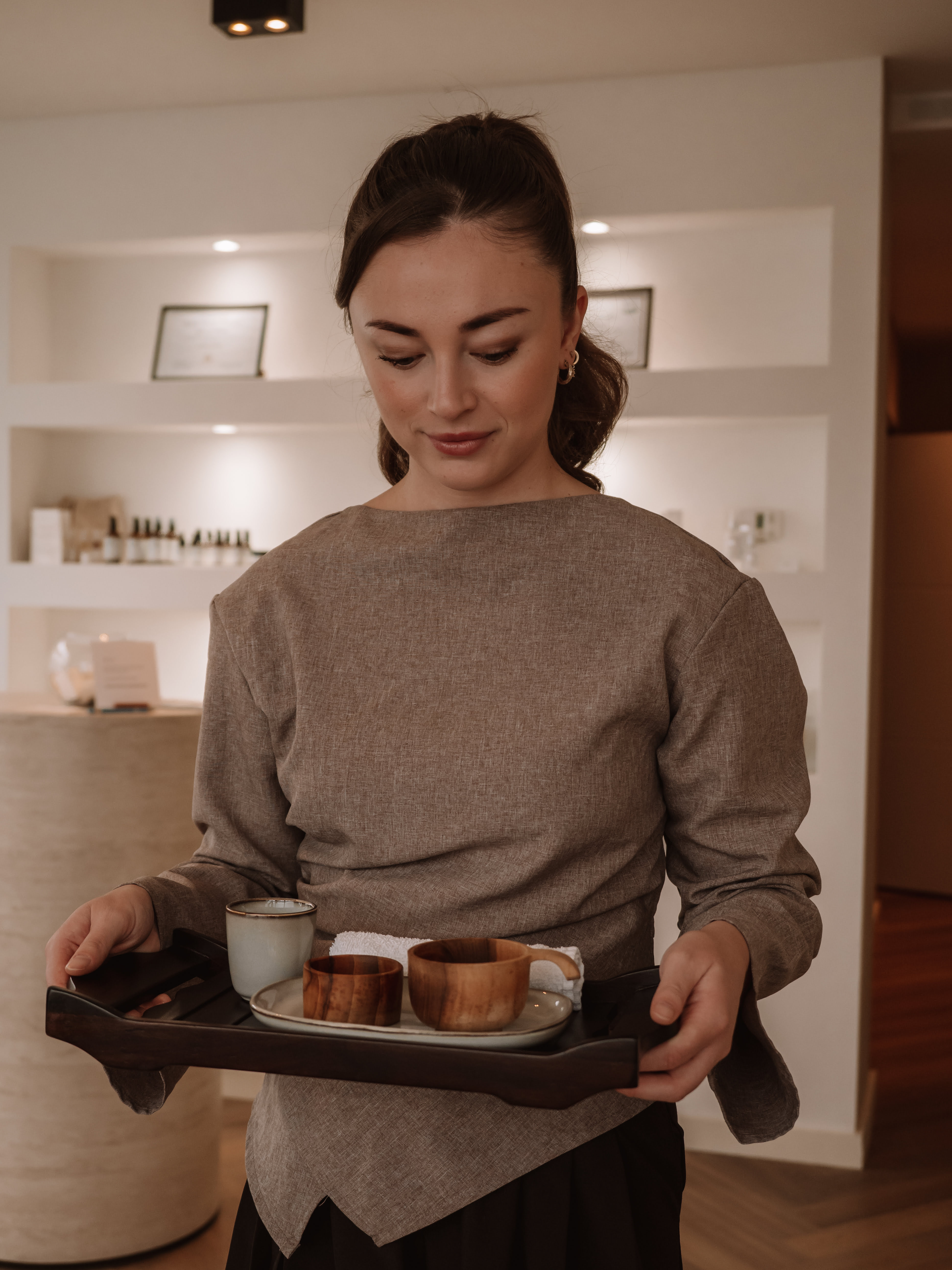 Woman in a brown long-sleeve top holding a wooden tray with ceramic and wooden cups, standing in a softly lit room.