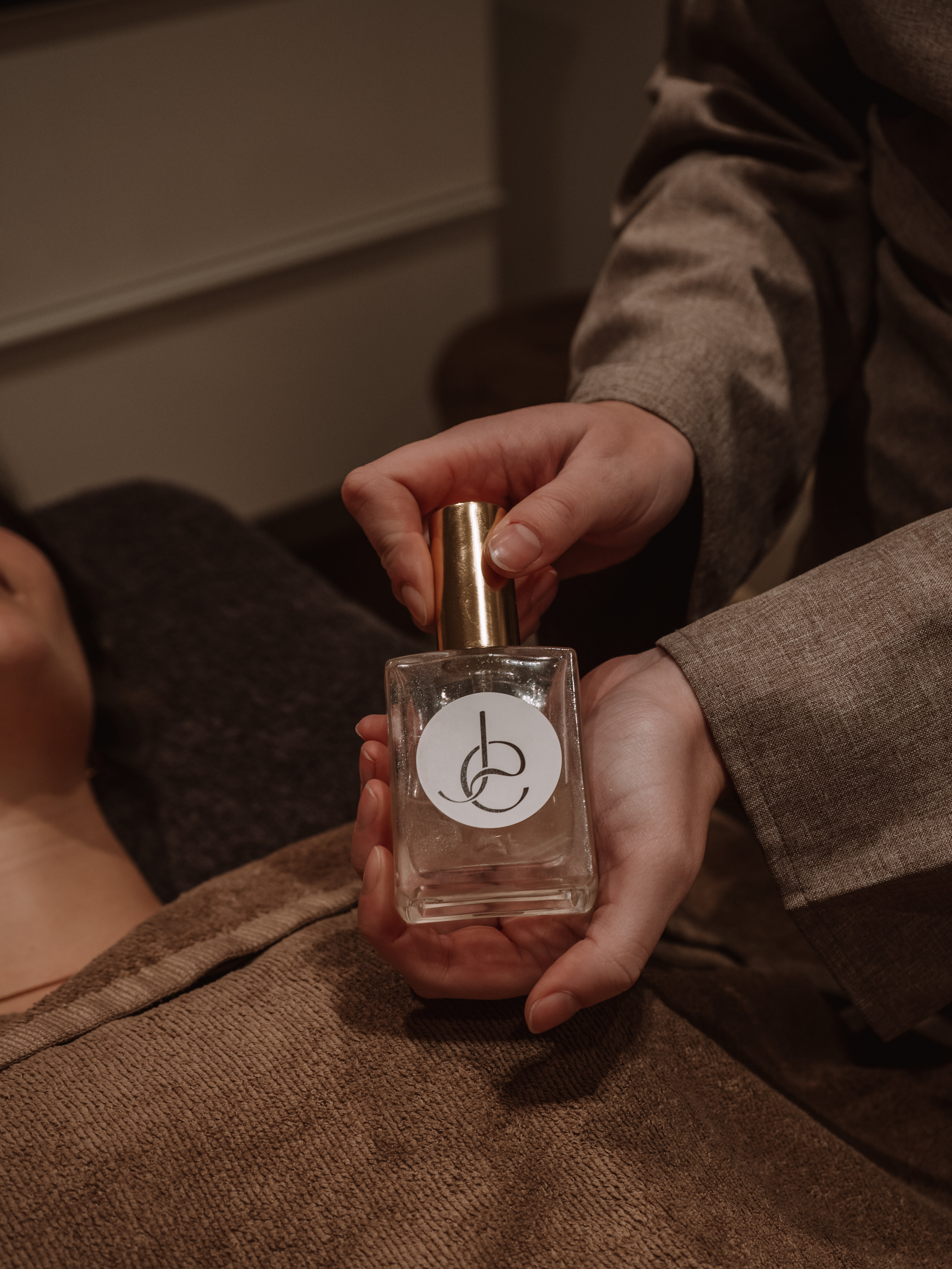 Hands holding a clear glass bottle with a gold cap, featuring a minimalist black logo on a white circle, above a person covered with a brown towel.