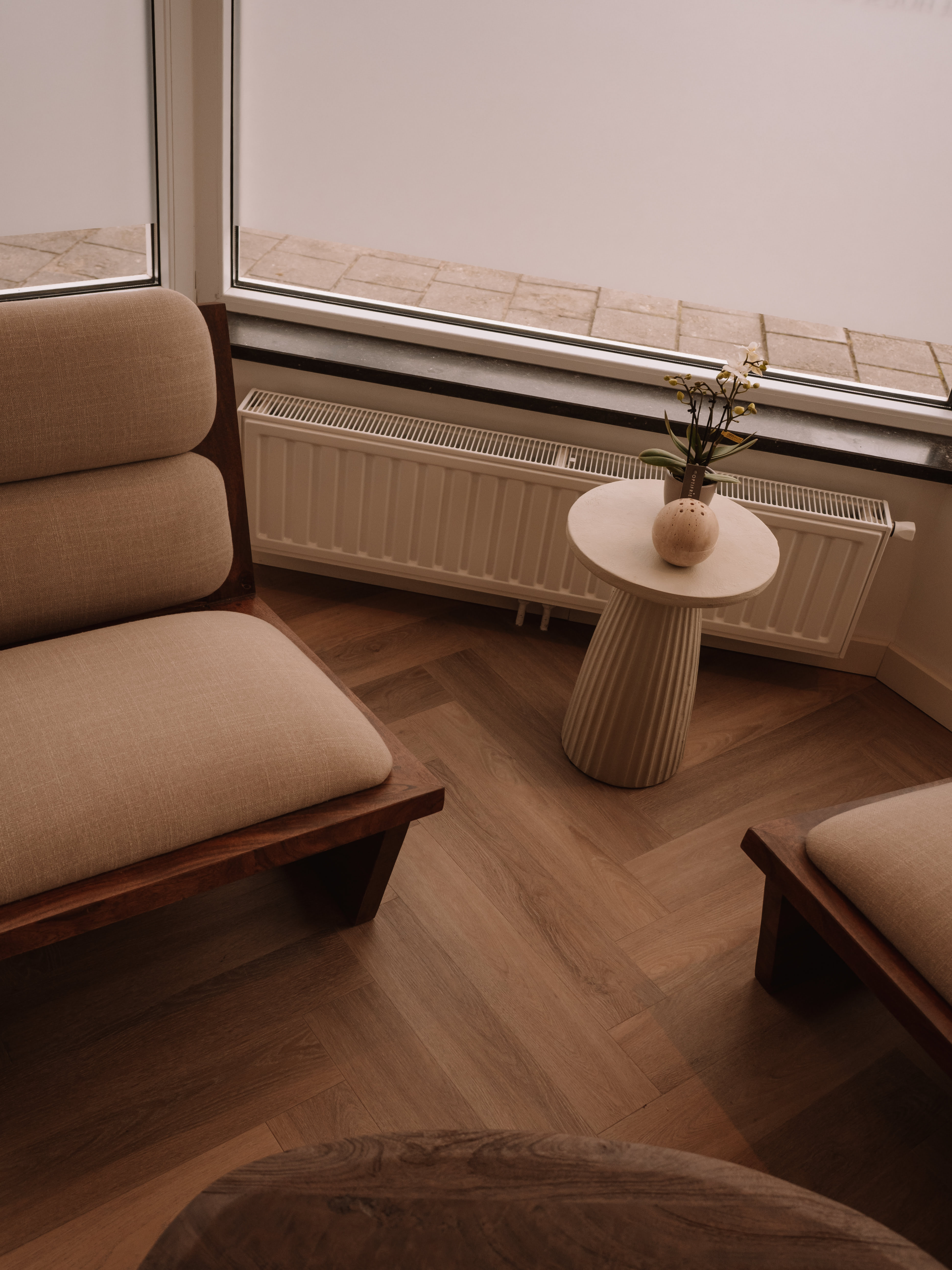 Cozy corner with two beige cushioned wooden chairs, a small round side table with a potted orchid and decorative item, set on herringbone wooden floor in front of a large window.