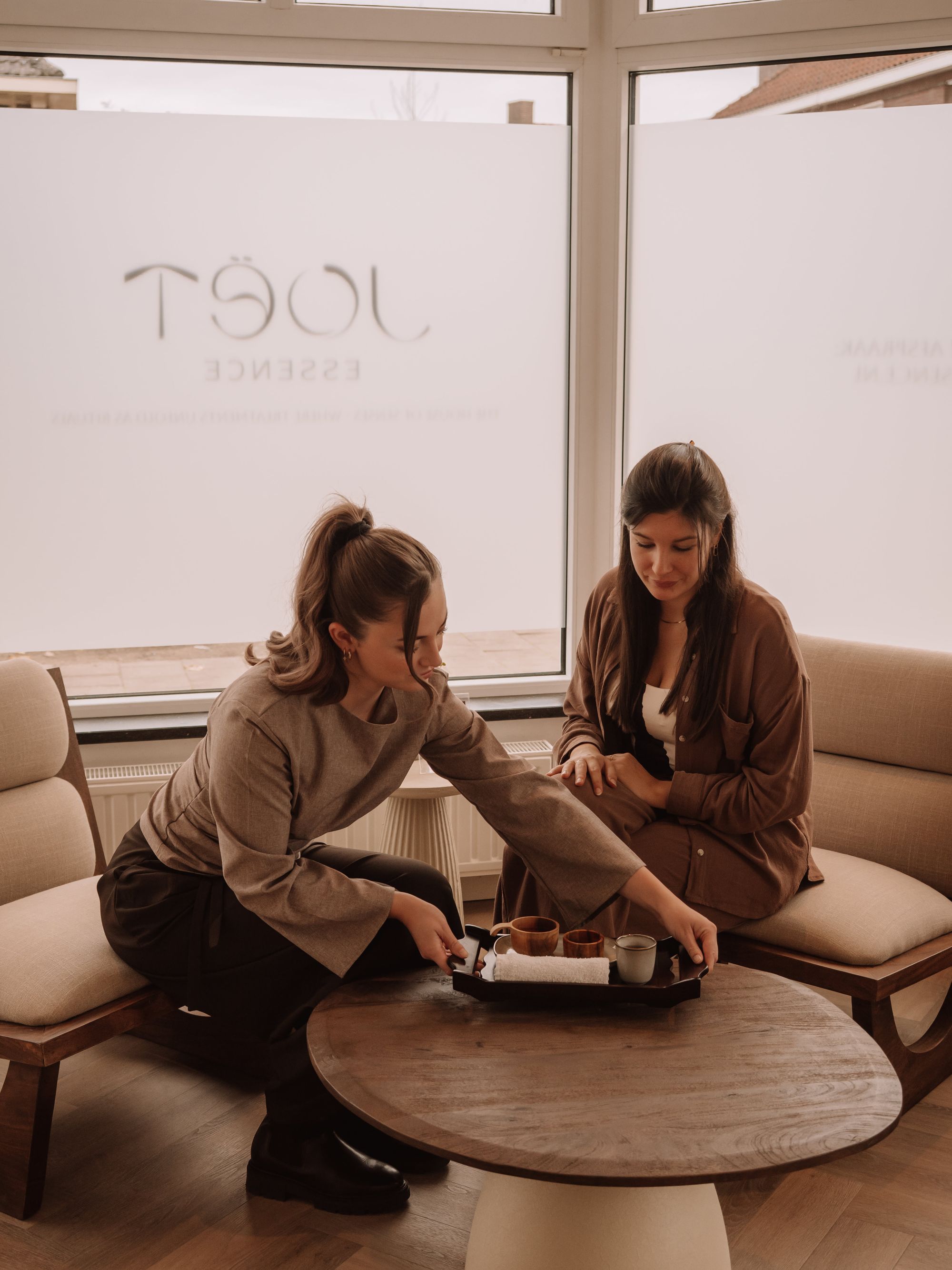 Two women sitting on beige chairs near a round wooden table, one placing a tray with cups and a towel on the table in a softly lit room.