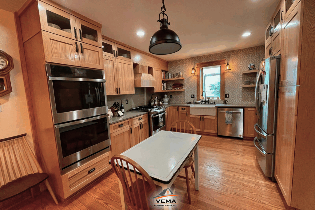 Modern kitchen featuring wooden cabinets, stainless steel double oven, refrigerator, dishwasher, farmhouse sink, wooden floor, and a white dining table with two wooden chairs under black pendant light.