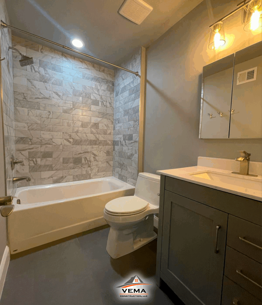 Modern bathroom with gray cabinetry, white bathtub and toilet, marble tile shower walls, and overhead lighting fixtures.