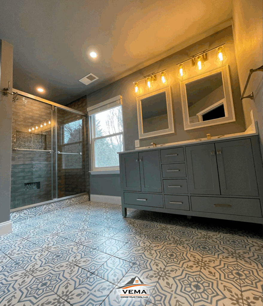 Modern bathroom with patterned tile floor, gray double vanity with two mirrors and exposed bulb lights, and a glass-enclosed shower with dark tiles.