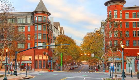 Urban street intersection lined with red brick buildings and trees with autumn foliage under a partly cloudy sky.