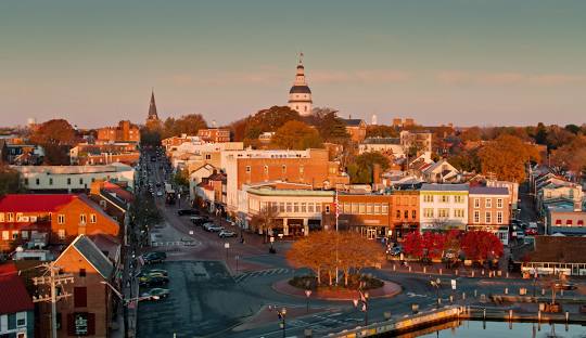 Aerial view of Annapolis, Maryland at sunset showing the cityscape, the Maryland State House dome, and a waterfront roundabout surrounded by autumn trees.