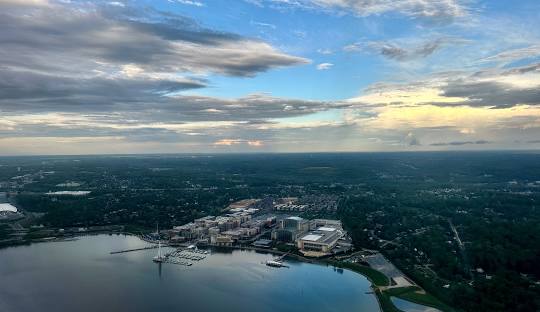 Aerial view of a waterfront cityscape with buildings, a marina, and a partly cloudy sky during sunset.