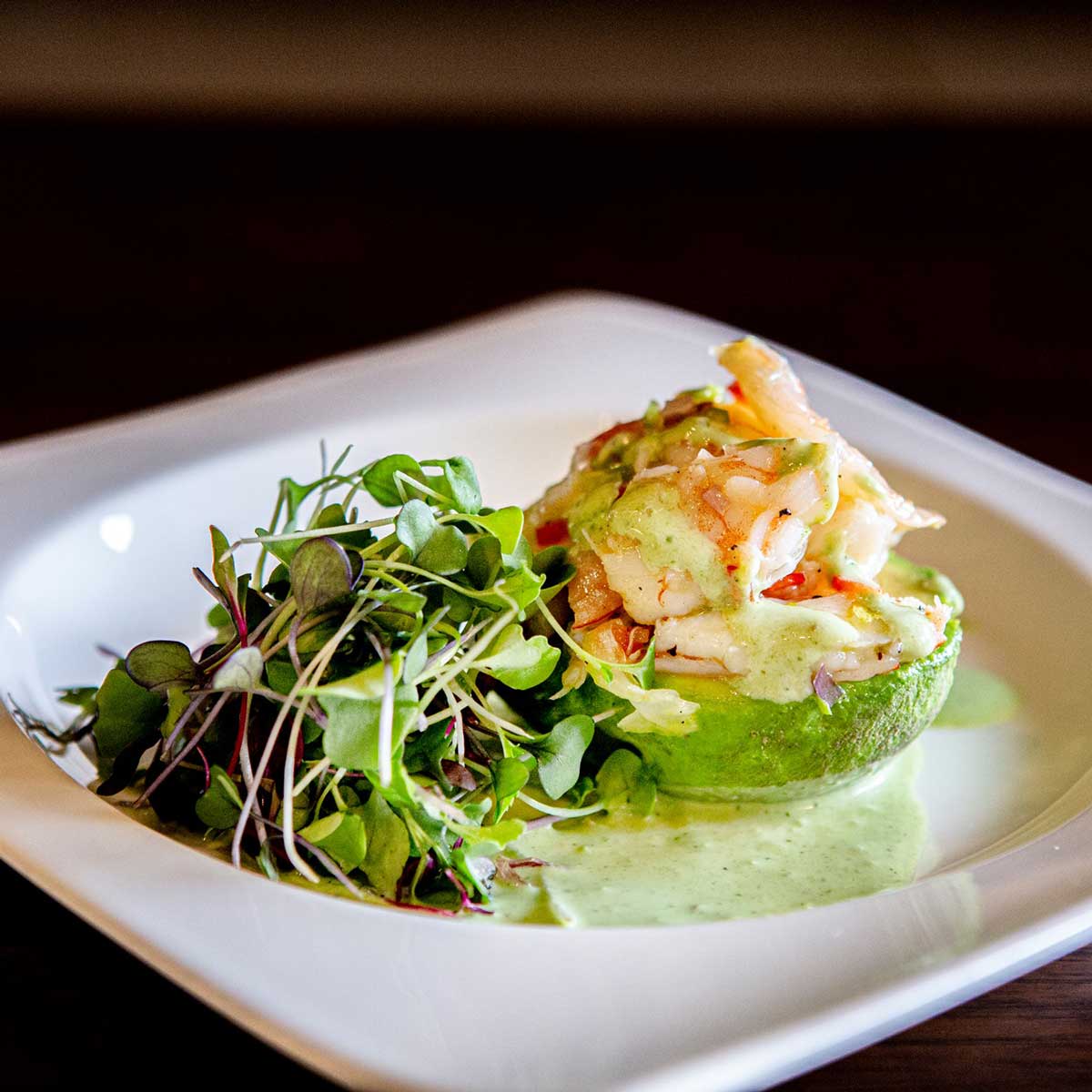 Grilled shrimp served on a halved avocado with green sauce and microgreens on a white plate.