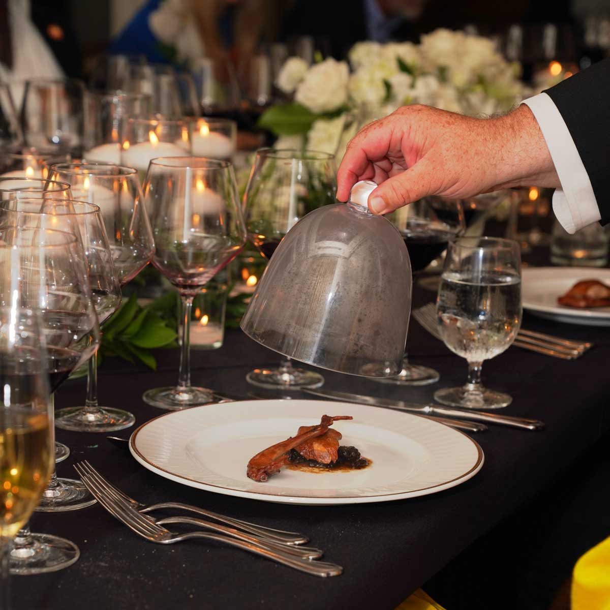 A hand lifting a glass cloche to reveal a gourmet dish of cooked bird on a white plate at a formal dining table with wine glasses and candles.