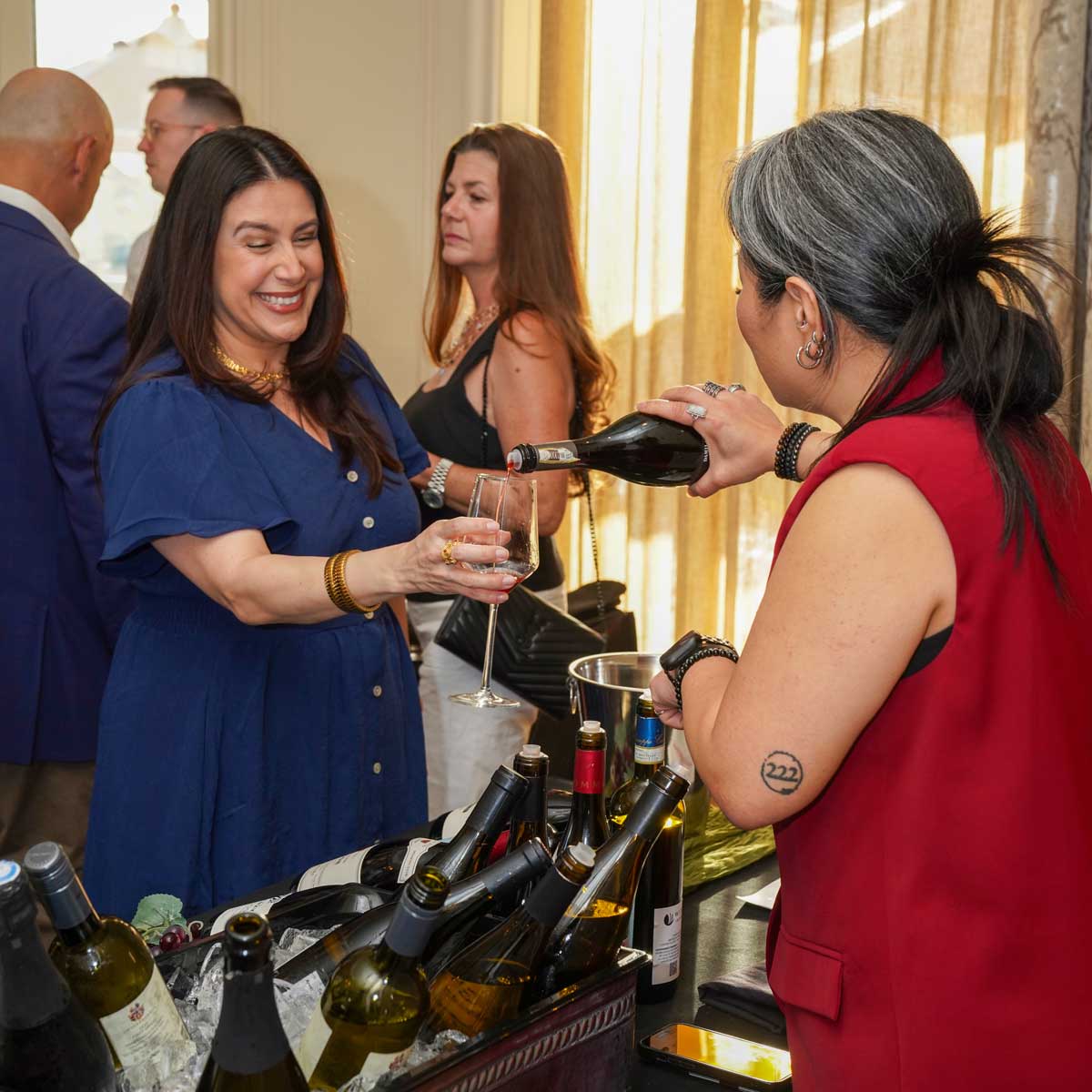 A woman in a red vest pours wine into a glass held by a smiling woman in a blue dress with other people in the background.
