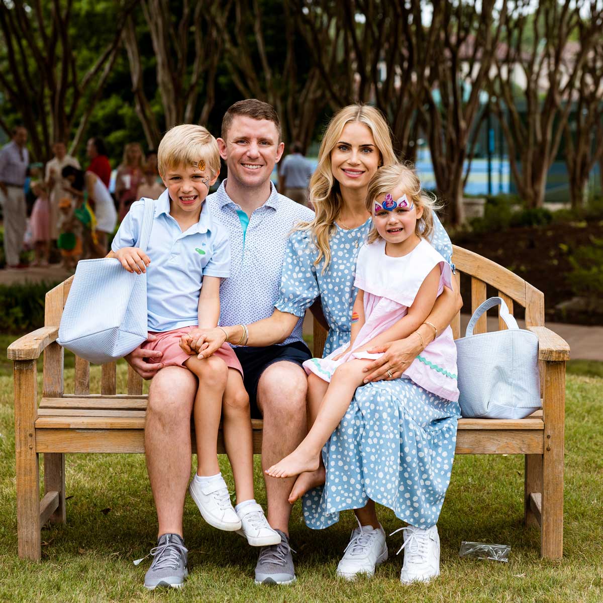 Family of four sitting on a wooden bench in a park, with the children having face paint and holding light blue bags.