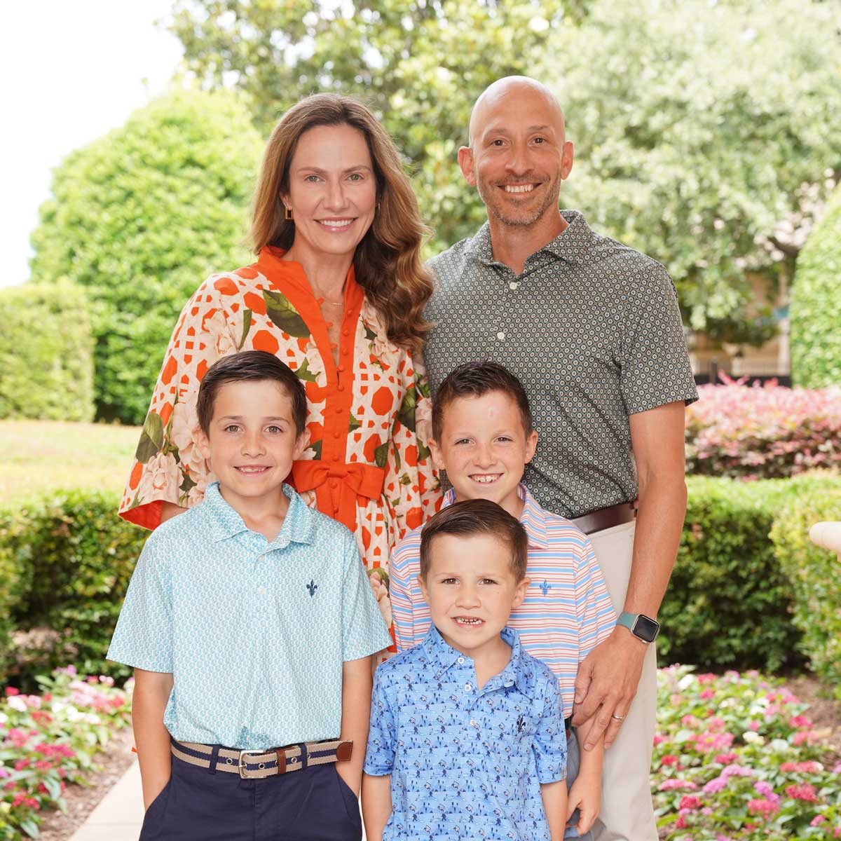 Smiling family of five posing outdoors in a garden with trimmed hedges and flowering plants.
