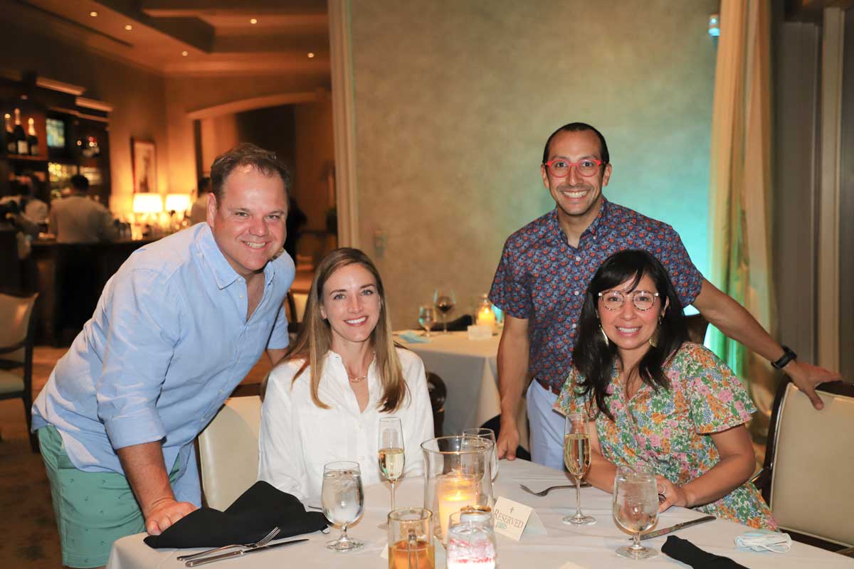 Four people smiling at a restaurant table set with glasses, cutlery, and a candle in a reserved spot.