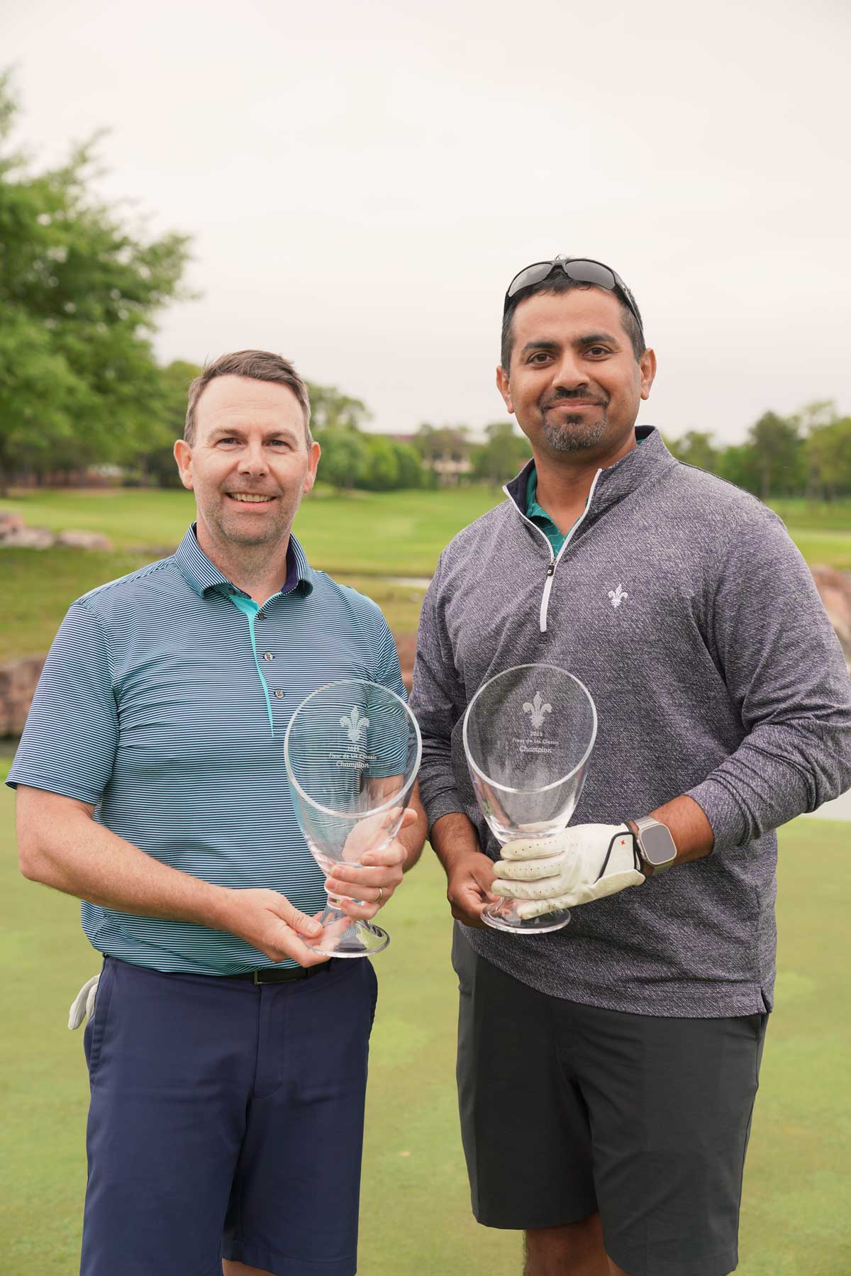 Two men standing on a golf course holding large glass trophies, smiling at the camera.