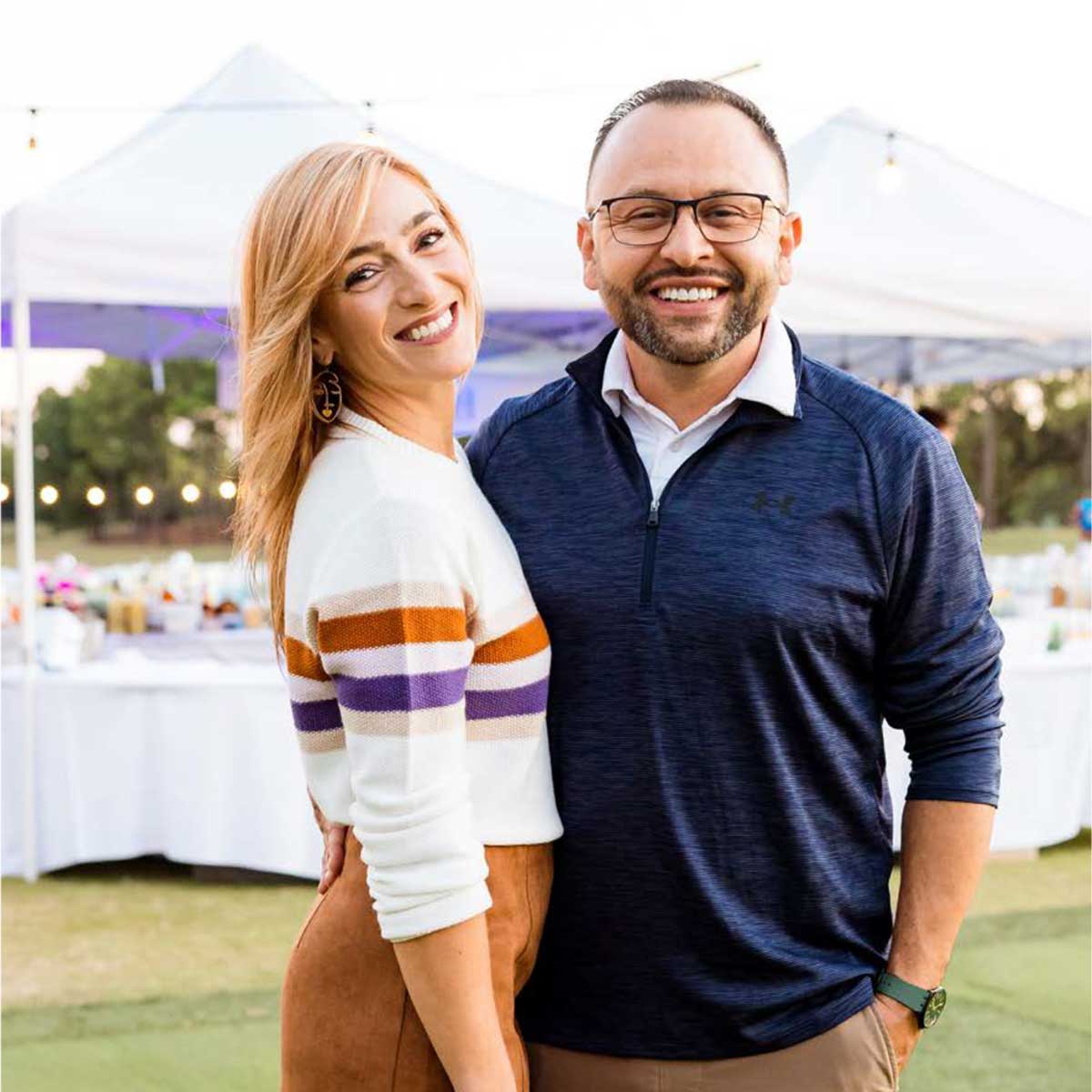 Smiling man with glasses and bearded face wearing a navy blue pullover and a smiling woman with blonde hair and striped sweater posing outdoors under white tents.