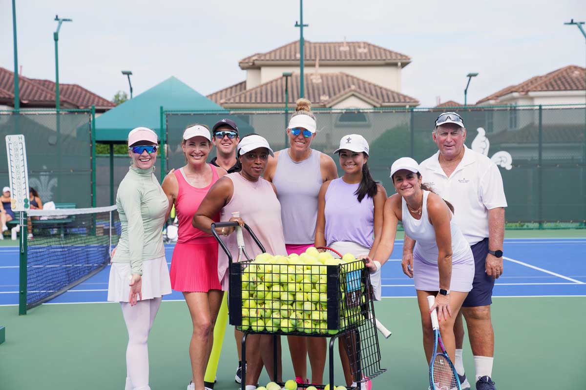 Group of seven tennis players standing on a court around a cart filled with tennis balls.