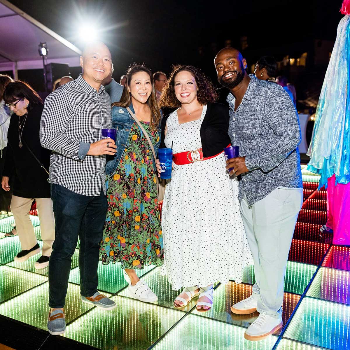 Four people smiling and holding drinks on a colorful illuminated floor at a nighttime event.