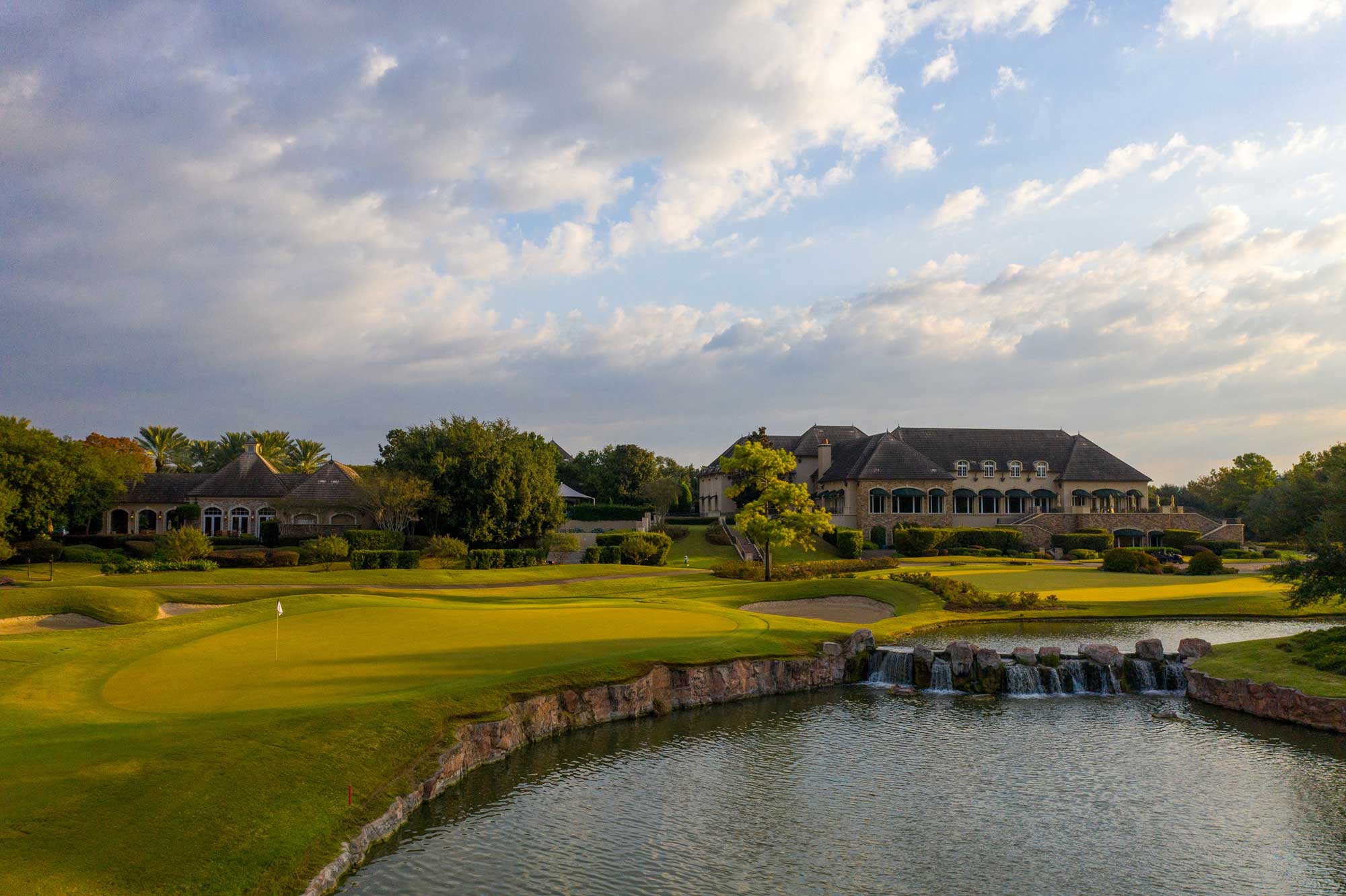Golf course green with a small waterfall and stone edge in front of a large clubhouse under a partly cloudy sky.