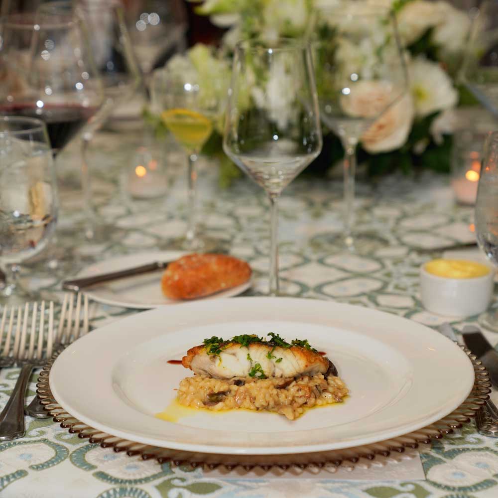 Plated dish of fish fillet garnished with herbs on a bed of risotto, served on an elegant table with wine glasses, bread roll, and floral centerpiece.