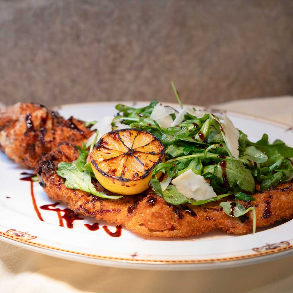 Breaded cutlet topped with arugula, shaved cheese, grilled lemon half, and drizzled with balsamic glaze on a white plate.