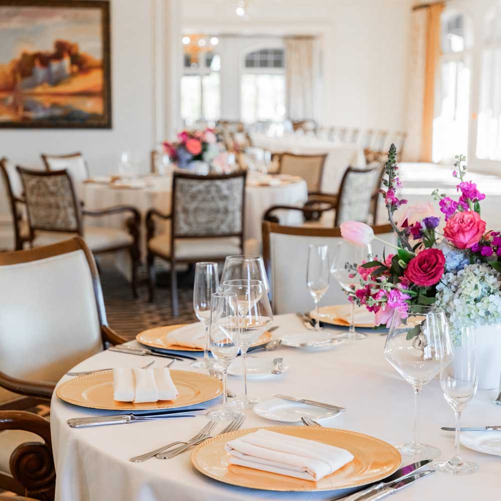 Elegant dining table set with gold plates, white napkins, crystal glassware, and a vibrant floral centerpiece in a bright, ornate room.