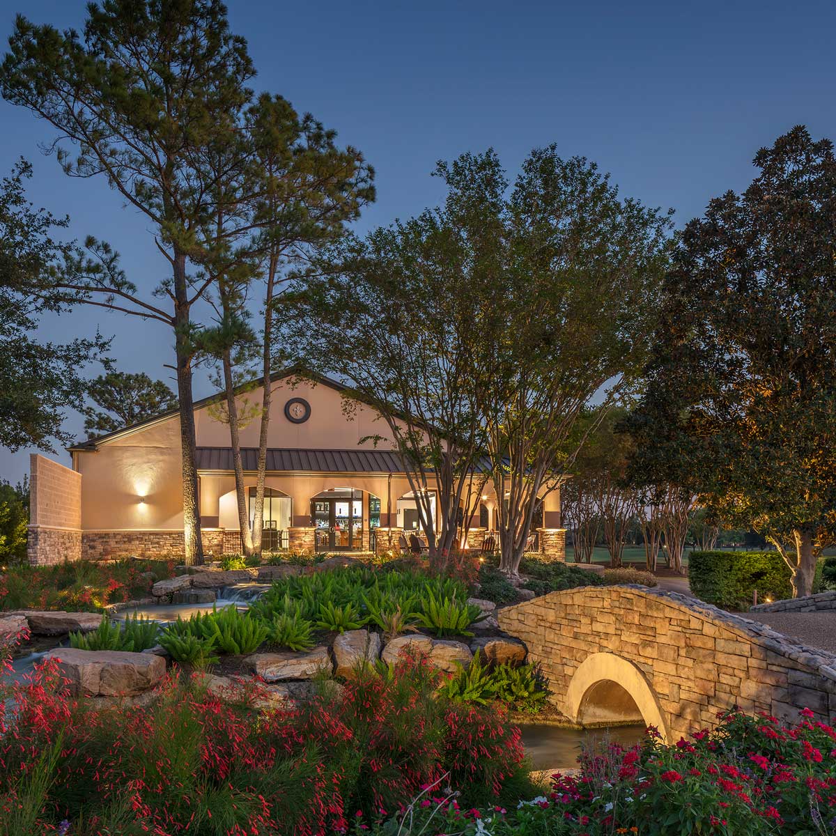 Evening view of a lit stone arch bridge over a landscaped garden pond with flowers, trees, and a beige building in the background.