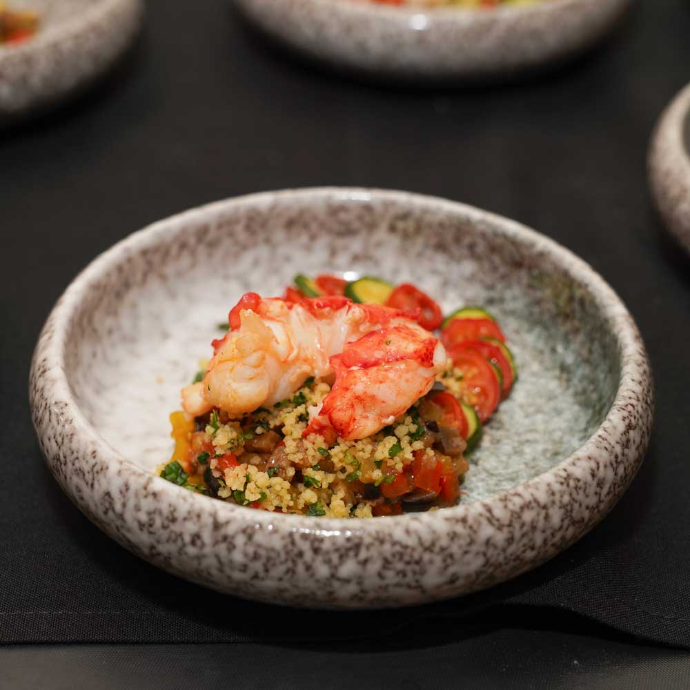 Cooked lobster tail served atop couscous with diced vegetables and garnished with sliced cherry tomatoes and zucchini on a speckled ceramic plate.
