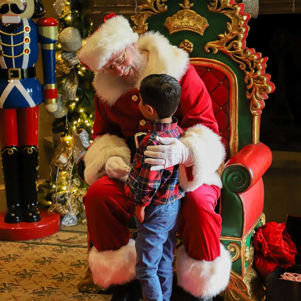 Santa Claus sitting on an ornate green and red throne, smiling and talking to a young boy wearing a plaid shirt and blue pants.