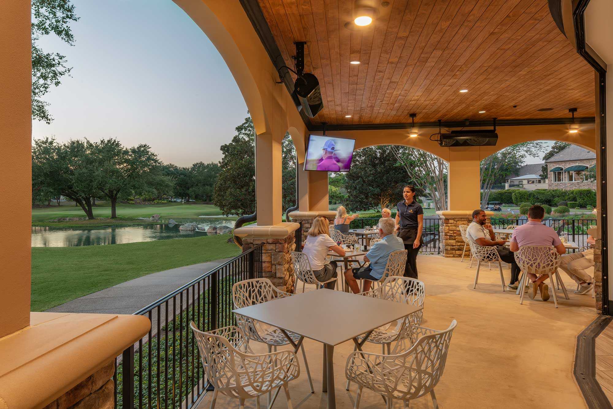 Outdoor covered patio with people sitting at tables overlooking a golf course pond and green landscape at dusk.