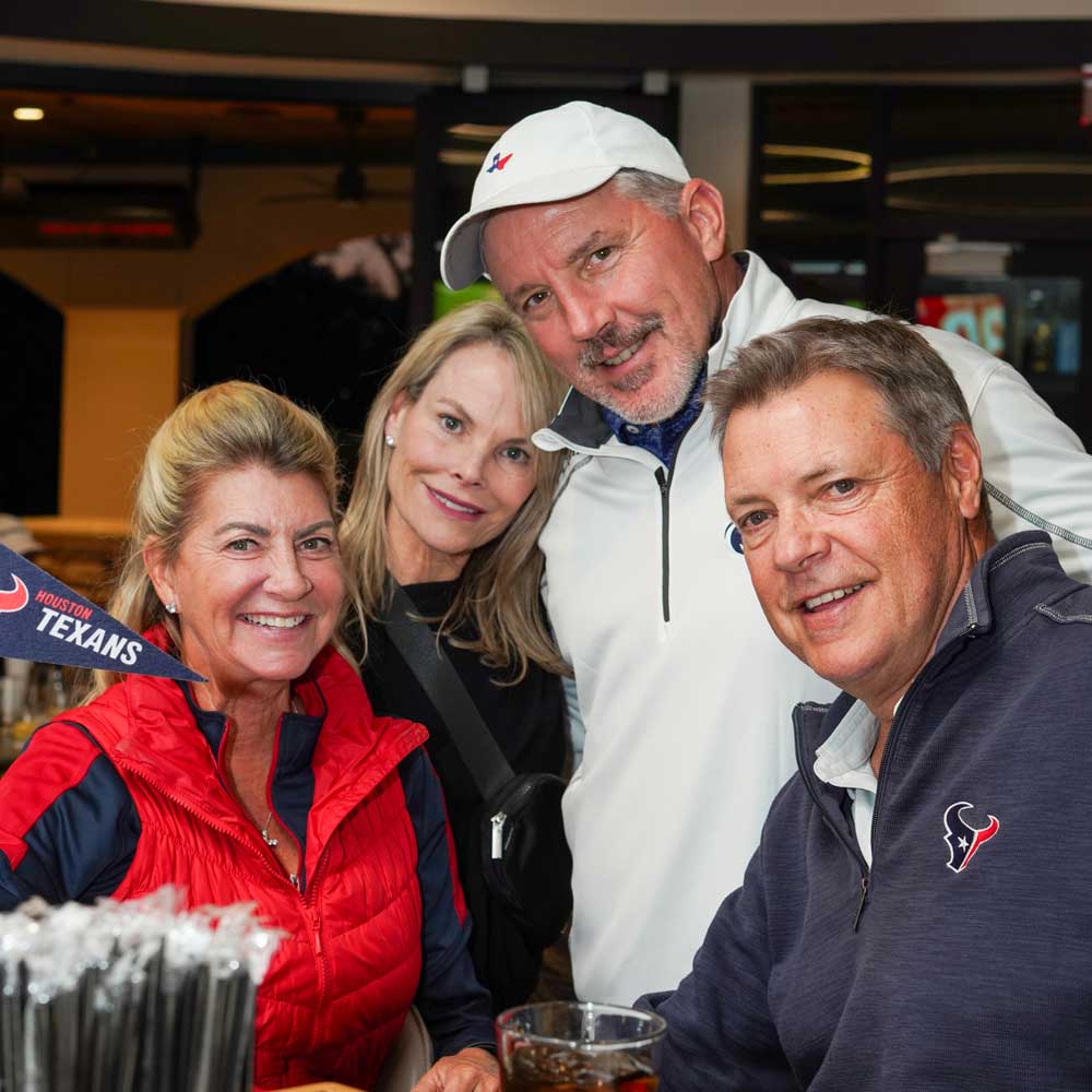 Four adults smiling indoors, with two wearing Houston Texans sportswear and a small Texans pennant visible.