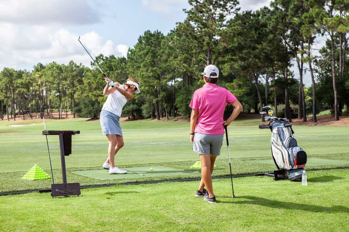 Two women at a golf driving range, one swinging a golf club and the other watching, surrounded by green grass and trees.