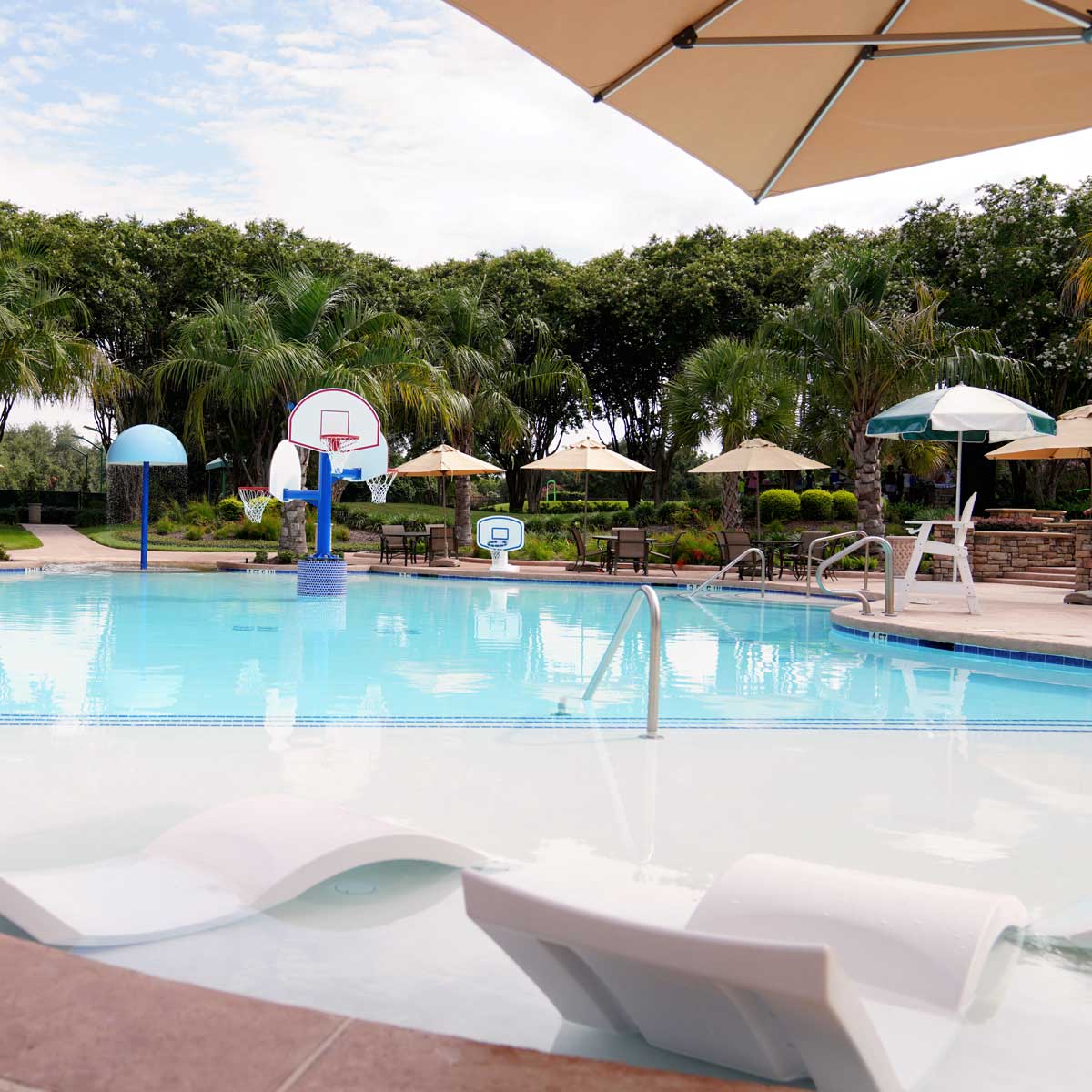 Outdoor swimming pool with built-in lounge chairs, basketball hoops, umbrellas, and surrounded by trees and lounge seating.