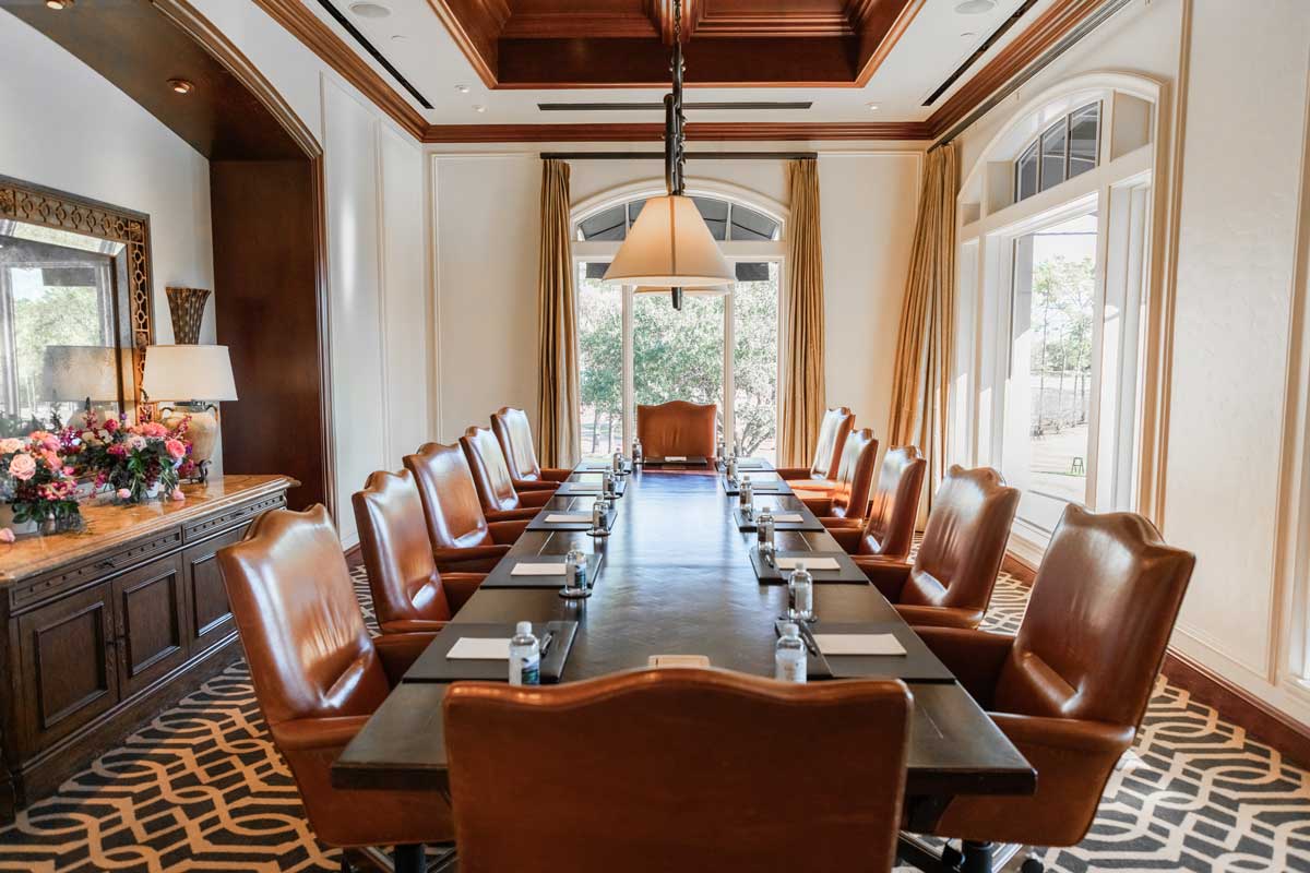 Elegant conference room with a long dark table surrounded by twelve leather chairs, windows with curtains, and a decorative sideboard with flowers and a lamp.