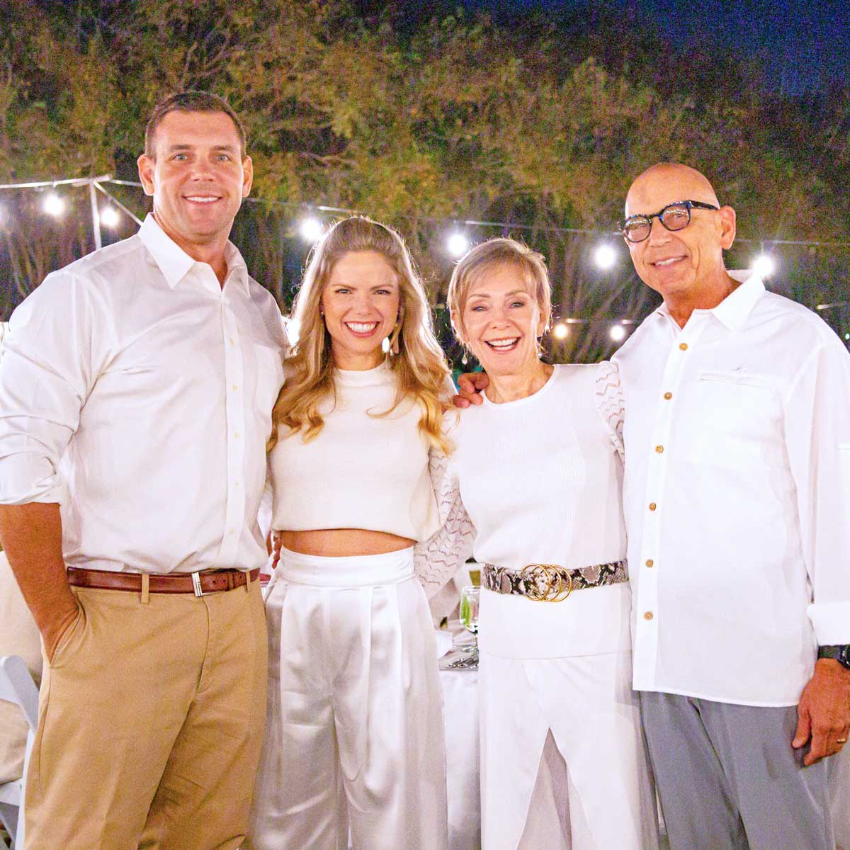 Four people dressed in white smiling and posing outdoors at a nighttime event with string lights in the background.