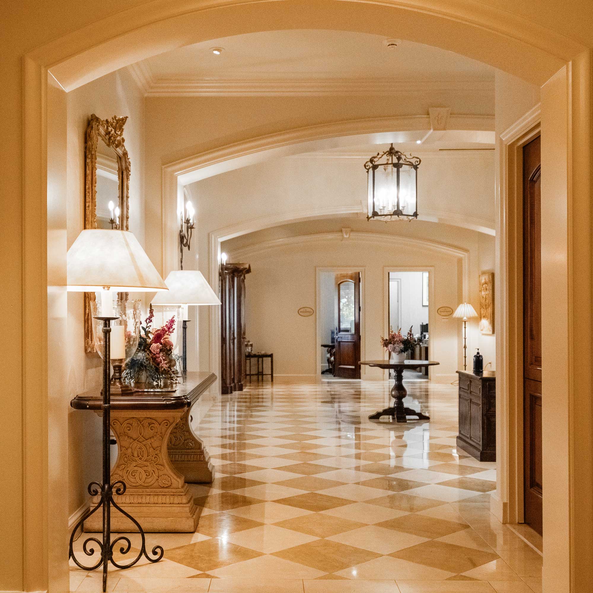 Elegant hallway with checkered floor, ornate console table with lamps and floral arrangement, and a round table with flowers under arched ceilings.